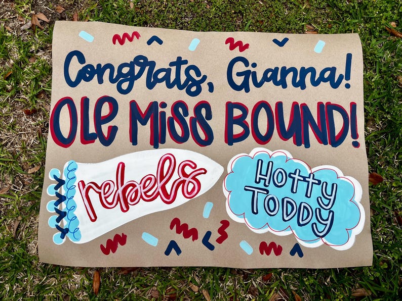 May include: A brown paper sign with the words "Congrats, Gianna! OLE MISS BOUND!" in blue and red lettering. The sign also features the word "rebels" in red and white, and "Hotty Toddy" in a blue cloud.