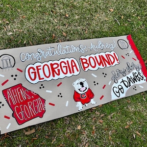 May include: A tan banner with red trim and the words "Congratulations, Audrey! GEORGIA BOUND!" in white and red. A bulldog in a red sweater and the words "Athens, Georgia" are also featured. The banner also says "Glory, Glory, Go Dawgs!"