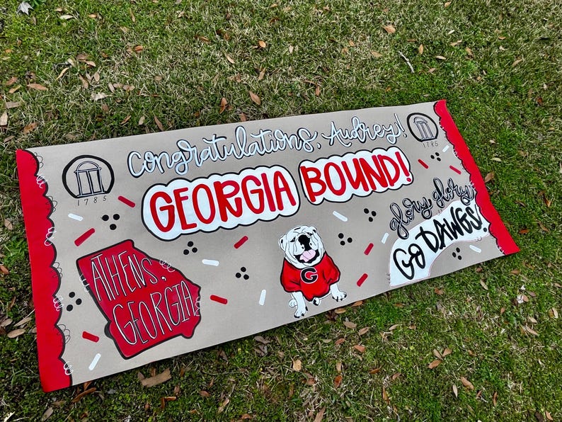 May include: A tan banner with red trim and the words "Congratulations, Audrey! GEORGIA BOUND!" in white and red. A bulldog in a red sweater and the words "Athens, Georgia" are also featured. The banner also says "Glory, Glory, Go Dawgs!"