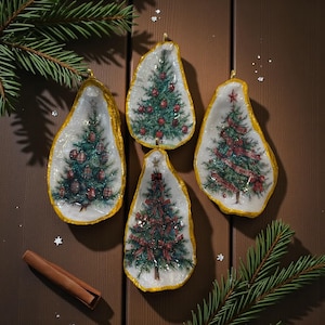 May include: Four Christmas tree ornaments in the shape of oyster shells. Each shell has a gold rim and a painted Christmas tree design. The trees are decorated with red ornaments and ribbons. The ornaments are displayed on a wooden surface with pine needles and a cinnamon stick.
