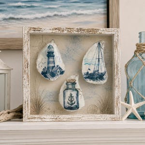 May include: A square, distressed white wooden shadow box frame containing three oyster shells painted with blue nautical imagery. The shells feature a lighthouse, a sailboat, and a glass bottle with an anchor, set against a sandy background.