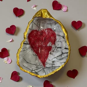 May include: A decorative oyster shell dish with a gold-painted rim and a red heart design in the center. The interior has a white and gray marbled pattern. Red heart-shaped petals are scattered around the dish.