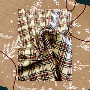 May include: A beige and red plaid fabric with a soft, woven texture. The fabric is folded and draped, showcasing the pattern and texture.