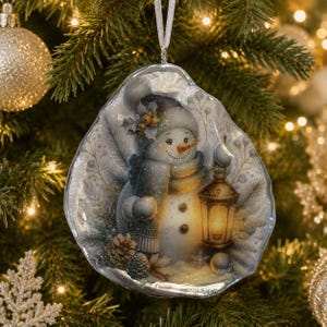 May include: A decorative oyster shell ornament featuring a painted snowman wearing a hat and scarf, holding a glowing lantern. The design includes pine branches and pinecones, finished with a silver ribbon for hanging on a Christmas tree.
