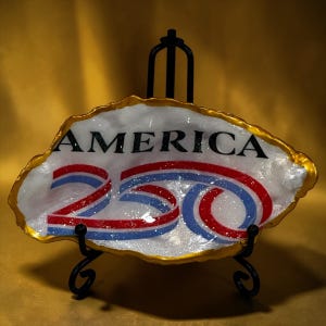 May include: A decorative, oval-shaped dish with the word "AMERICA" in black and the number "250" in red, white, and blue. The dish has a gold-colored rim and sits on a black metal stand. The background is a solid gold color.