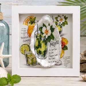 May include: A square white frame containing a print of cocktail recipes including Tequila Sunrise, Mojito, and Pina Colada. A large, iridescent oyster shell is mounted in the center, featuring a hand-painted illustration of a Mojito with mint.