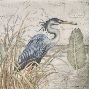May include: A decorative paper napkin featuring a detailed illustration of a blue heron standing in tall grass. The background includes a vintage map design with the words "Caribbean Sea" and "Atlantic". The heron has a long beak and is surrounded by foliage.