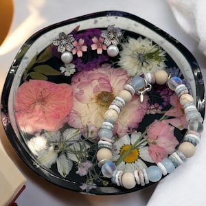 May include: A black resin tray with a floral design featuring pressed flowers and leaves. The tray also includes a pair of silver earrings with pearl accents and a beaded bracelet with white and blue beads.