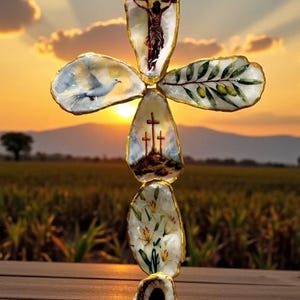 May include: A handcrafted cross made from oyster shells, featuring painted religious imagery. The cross includes depictions of Jesus on the cross, a dove, olive branches, and lilies. The shells are edged with gold and set against a sunset backdrop.