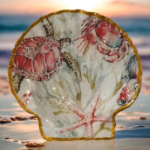 May include: A decorative seashell-shaped dish with a gold rim. The interior features watercolor illustrations of a red sea turtle, crab, starfish, and fish, with seaweed accents. The background is a beach scene.
