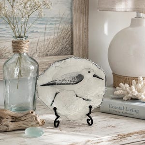 May include: A circular glass plate with a textured, translucent white finish featuring a black and white illustration of a sandpiper bird in profile. The plate is supported by a black metal display stand with curved feet and a decorative edge.