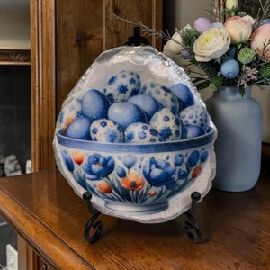 May include: A decorative bowl filled with blue Easter eggs, resting on a black metal stand. The bowl features a floral design with blue and orange flowers. The eggs are decorated with white floral patterns.