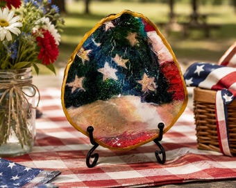 May include: A decorative, shell-shaped dish featuring a painted American flag design with white stars on a blue background and red and white stripes. The dish is displayed on a black metal stand, set on a red and white checkered tablecloth, with a picnic basket and flowers in the background.