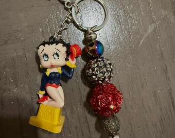 Puede incluir: Un llavero con una figura de Betty Boop con un atuendo azul y amarillo, con sombrero y zapatos rojos. El llavero también incluye un dije con cuentas plateadas, rojas e iridiscentes. La base de la figura dice "USA".