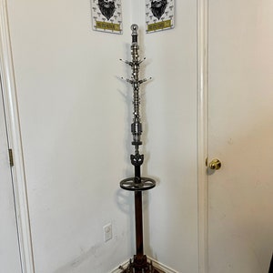 May include: A coat rack made from repurposed car parts. The rack is made of metal and has a silver finish. The rack has a round base and a tall, slender shaft. The shaft is decorated with various car parts, including a crankshaft, pistons, and gears. The rack has a round shelf at the top for hanging coats. The rack is standing in a corner of a room with white walls and a white door. Two signs are hanging on the wall behind the rack. The signs are white with black text and images. The text on the signs reads 