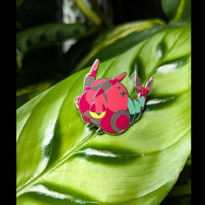 Peut inclure: Épingle en émail colorée représentant une créature stylisée. L'épingle a un corps rouge avec des accents gris et roses, un œil jaune et des sections turquoise. Elle est posée sur une feuille verte.