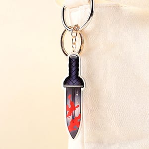 May include: A keychain featuring a dagger design. The dagger has a black handle with a woven pattern and a blade with red splatter details. The keychain is attached to a silver keyring and a gold-colored ring.