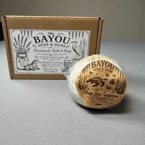 May include: A brown cardboard box and a round bath bomb. The box has a label with the text "BAYOU SUDS & DUDS Handmade Bath & Body" and illustrations. The bath bomb is wrapped and has a label that reads "Deep Sea Surprise".