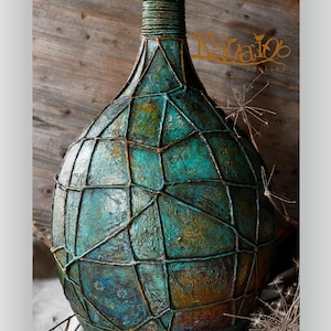 May include: A decorative vase with a textured teal and bronze finish, overlaid with a geometric wire pattern. The neck is a darker green. The vase has a rustic, artistic design. The height of the vase is approximately 40 cm.