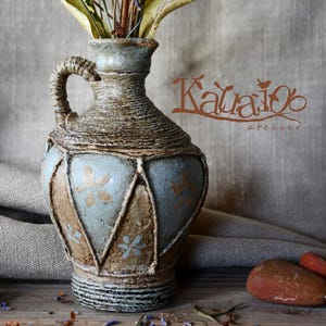 May include: A rustic vase with a handle, holding dried flowers and foliage. The vase has a blue and brown colour scheme, with a textured, rope-like pattern. The text "Kataios atelier" is visible on the right side.