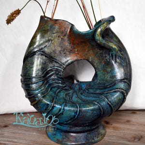 May include: A handcrafted ceramic vase with an abstract design. The vase has a blend of teal, bronze, and copper colours, with a textured surface. Dried grasses and stems are arranged inside. The artist's name, Katia06, is visible.