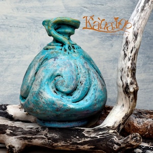 May include: A turquoise ceramic vase with a textured, swirling relief pattern and a weathered finish. The vase features a narrow neck with a decorative coiled handle element. The text "Kala100" is visible in the upper right corner of the image.