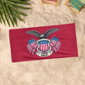 May include: A red beach towel featuring a patriotic design. The design includes a bald eagle with outstretched wings, a shield with stars and stripes, and American flags. The name "ASHLEY" is displayed on a banner.