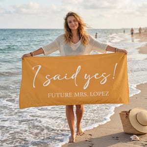May include: Mustard yellow beach towel with the text "I said yes!" and "Future Mrs. Lopez" in white script. The towel is held up by a person on a sandy beach with the ocean in the background.