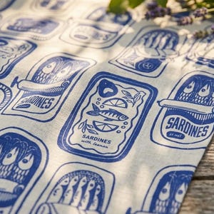K&ouml;nnte beinhalten: Ein wei&szlig;es Leinentuch mit einem sich wiederholenden blauen Druck von Sardinenb&uuml;chsen. Die Illustrationen zeigen verschiedene Sardinenb&uuml;chsen-Designs mit Text, darunter "Sardines with lemon" und "Saisines". Der Stoff liegt auf einer Holzoberfl&auml;che.