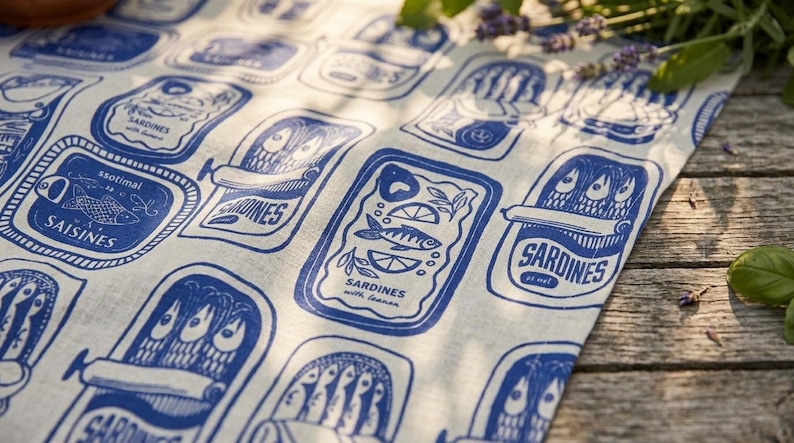 K&ouml;nnte beinhalten: Ein wei&szlig;es Leinentuch mit einem sich wiederholenden blauen Druck von Sardinenb&uuml;chsen. Die Illustrationen zeigen verschiedene Sardinenb&uuml;chsen-Designs mit Text, darunter "Sardines with lemon" und "Saisines". Der Stoff liegt auf einer Holzoberfl&auml;che.