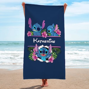 May include: A navy blue beach towel with a personalized name "Katherine" printed in white. The towel features a design of three blue Stitch characters from the Disney movie Lilo & Stitch, surrounded by pink flowers and green leaves.