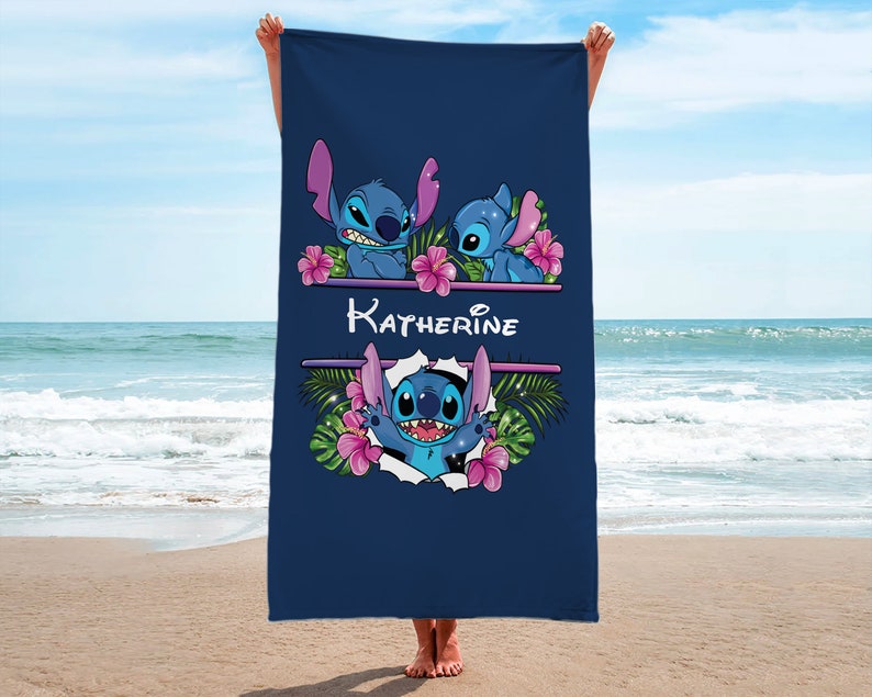 May include: A navy blue beach towel with a personalized name "Katherine" printed in white. The towel features a design of three blue Stitch characters from the Disney movie Lilo & Stitch, surrounded by pink flowers and green leaves.