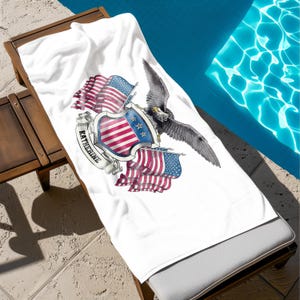 May include: White beach towel with a patriotic design featuring the American flag, an eagle, and the name "Katherine" on a banner. The towel is draped over a wooden lounge chair next to a pool.