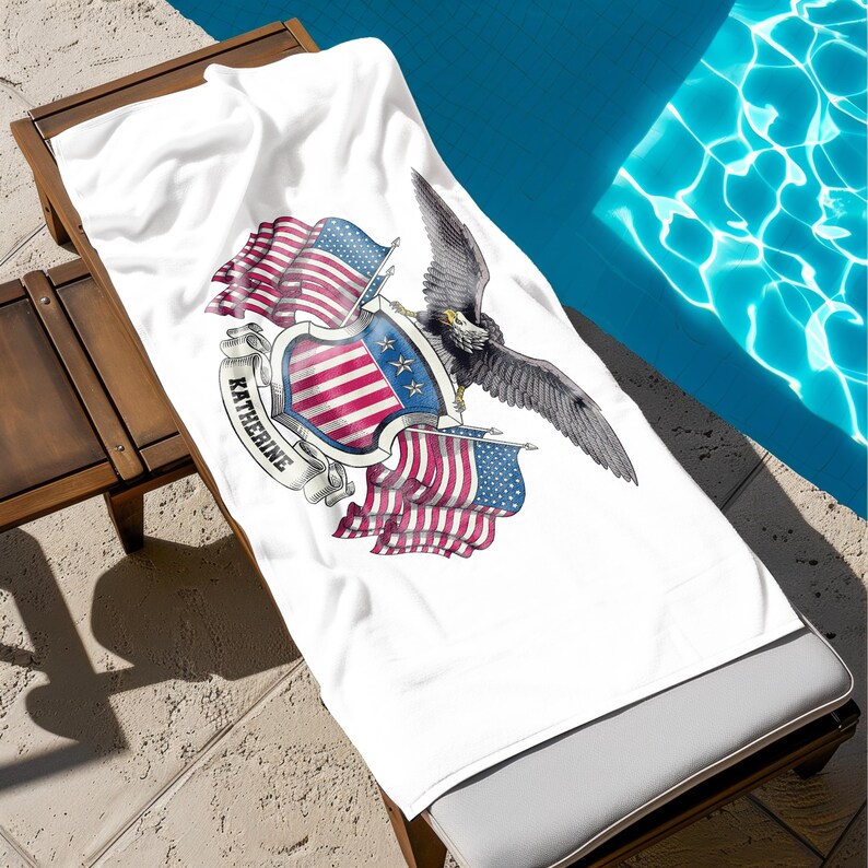 May include: White beach towel with a patriotic design featuring the American flag, an eagle, and the name "Katherine" on a banner. The towel is draped over a wooden lounge chair next to a pool.