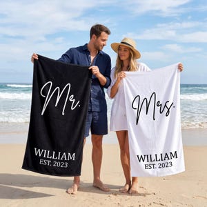 May include: A black beach towel with the word "Mr." in white script, the name "WILLIAM" and the year "EST. 2023". A white beach towel with "Mrs." in script, the name "WILLIAM" and the year "EST. 2023".