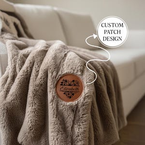 May include: A plush, taupe-colored blanket with a custom patch design. The round, brown leather patch features floral details and the name "Elizabeth" in elegant script. The text "CUSTOM PATCH DESIGN" is also visible.