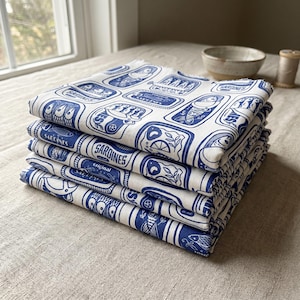 K&ouml;nnte beinhalten: Ein Stapel wei&szlig;er Stoffservietten mit einem blauen Sardinenb&uuml;chsen-Design. Jede Serviette zeigt ein sich wiederholendes Muster von Sardinenb&uuml;chsen-Illustrationen mit dem Wort "Sardines" und anderem Text. Die Servietten sind ordentlich gefaltet und auf einer neutralfarbenen Oberfl&auml;che gestapelt.