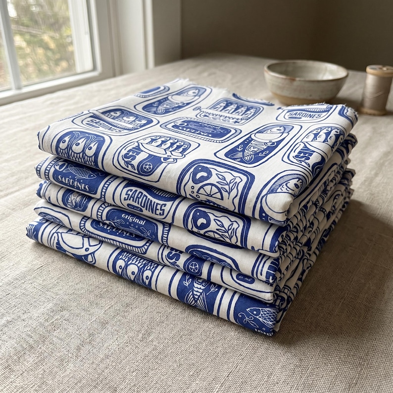 K&ouml;nnte beinhalten: Ein Stapel wei&szlig;er Stoffservietten mit einem blauen Sardinenb&uuml;chsen-Design. Jede Serviette zeigt ein sich wiederholendes Muster von Sardinenb&uuml;chsen-Illustrationen mit dem Wort "Sardines" und anderem Text. Die Servietten sind ordentlich gefaltet und auf einer neutralfarbenen Oberfl&auml;che gestapelt.
