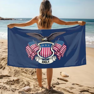 May include: A navy blue beach towel featuring a patriotic design with an eagle, American flags, and a shield. The towel has the name "EMILY" on a banner. The person is standing on a sandy beach with the ocean in the background.