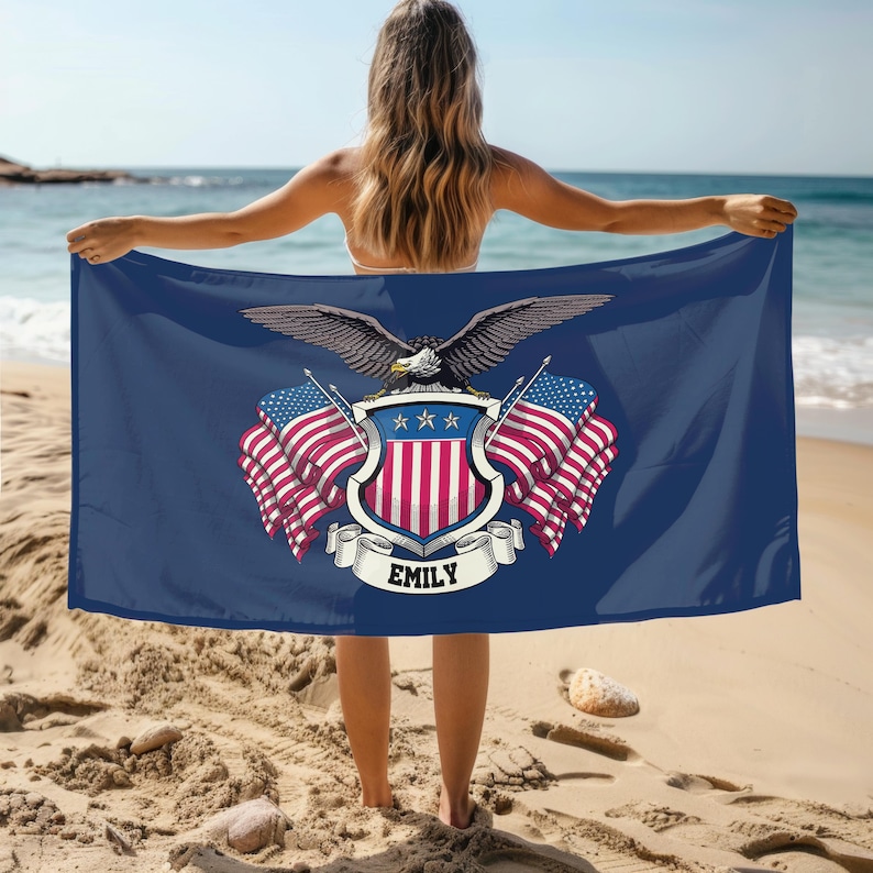 May include: A navy blue beach towel featuring a patriotic design with an eagle, American flags, and a shield. The towel has the name "EMILY" on a banner. The person is standing on a sandy beach with the ocean in the background.