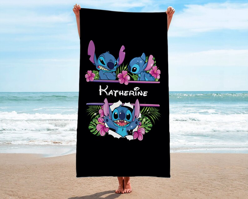 May include: Black beach towel with a personalized name "Katherine" and a blue Stitch character from Disney's Lilo & Stitch. The Stitch character is surrounded by pink flowers and green leaves.