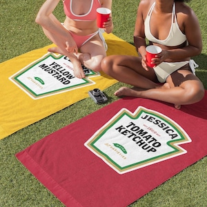 May include: Two beach towels with a ketchup and mustard design. The towels are yellow and red with white text and green accents. The text on the towels reads "Yellow Mustard" and "Tomato Ketchup".