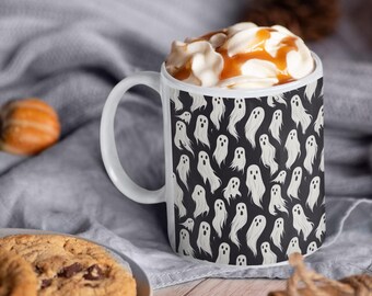 May include: White ceramic mug with a black background covered in white ghost illustrations. The mug is filled with whipped cream and caramel sauce. A chocolate chip cookie and cinnamon sticks are also in the image.
