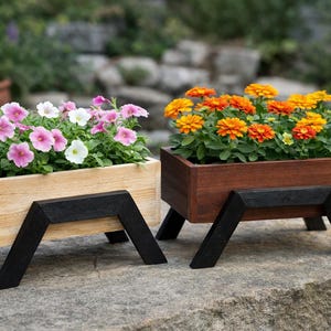 Op de afbeelding: Twee houten plantenbakken met bloemen. Eén plantenbak is lichtgekleurd en bevat roze en witte petunia's. De andere is donkerbruin en gevuld met oranje en gele zinnia's. Beide plantenbakken hebben zwarte schuine poten.