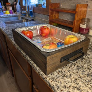 May include: A wooden serving tray with a metal handle holds a disposable aluminum pan containing several red and yellow apples. The tray sits on a granite countertop in a kitchen setting.