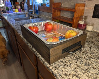 Op de afbeelding: Een houten serveerblad met een metalen handvat bevat een wegwerp aluminium pan met verschillende rode en gele appels. De lade staat op een granieten aanrecht in een keuken.