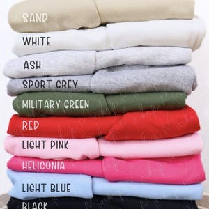 May include: A stack of folded sweatshirts in various colors, including sand, white, ash, sport grey, military green, red, light pink, heliconia, light blue, and black. Each sweatshirt has a label indicating its color. The sweatshirts are neatly arranged, showcasing a range of hues.