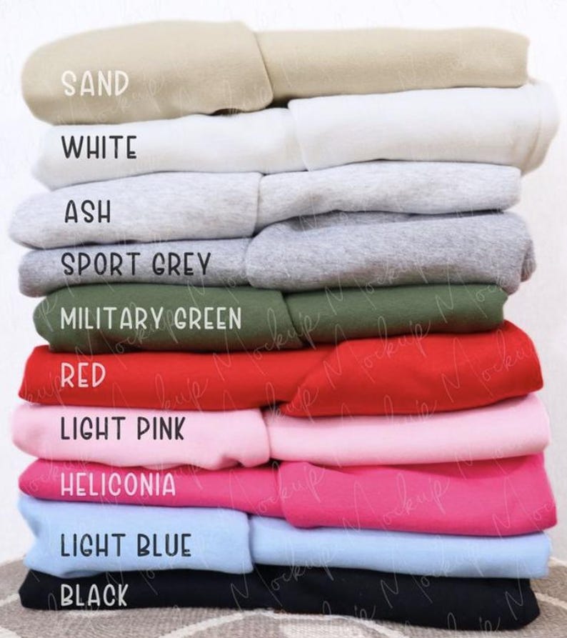 May include: A stack of folded sweatshirts in various colors, including sand, white, ash, sport grey, military green, red, light pink, heliconia, light blue, and black. Each sweatshirt has a label indicating its color. The sweatshirts are neatly arranged, showcasing a range of hues.
