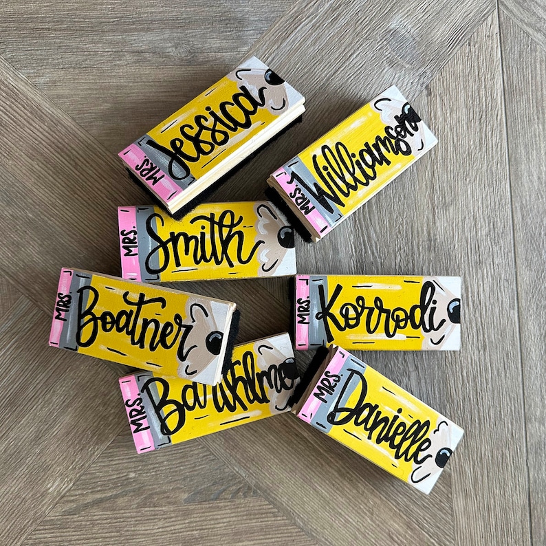 May include: Nine yellow and black pencil-shaped wooden signs with the text "Mrs." followed by a teacher's last name. The signs are painted to look like pencils with a pink eraser on the top.