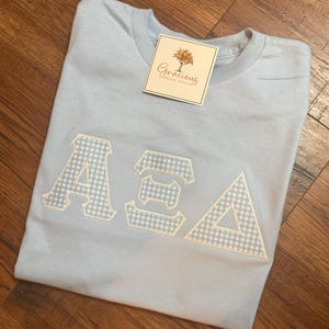 May include: Light blue t-shirt with the Greek letters ASA in white and light blue gingham fabric. The shirt has a tag that reads "Gracious Grove Studio". The shirt is folded on a wooden surface.